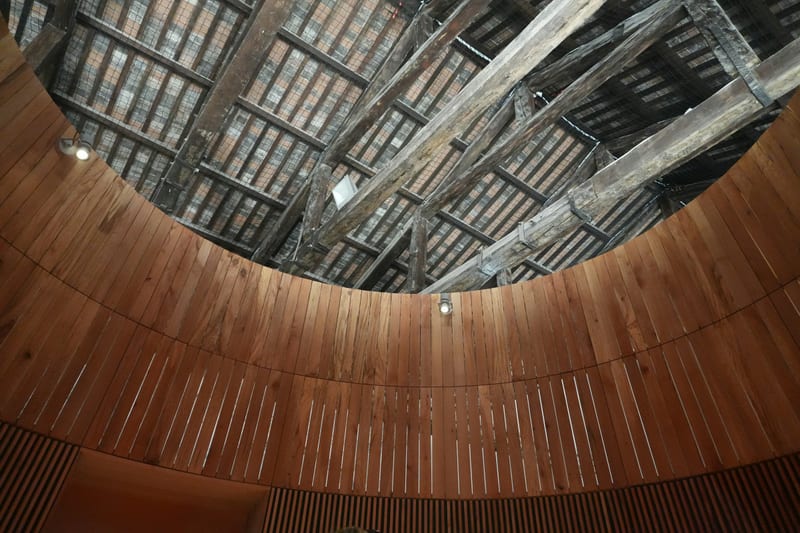 Korunmus eski ahsap cati kirisleri/makaslarinin altina yerlestirilmis yeni kavisli ahsap lameli hacim; yeni gomme spotlar kirislerin altinda. Adaptive reuse'un ozeti: eski tasiyici acikta, yeni eleman icine oturtulmus. Insan yok, net, ahsap ve toprak tonlari.