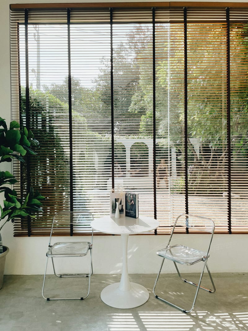 Pencere onunde iki minimal sandalye ve kucuk masa; minimalist kafe ic mekani. Insan yok, tabela/logo yok. Dogal gunisigi pencereden giriyor, gri-notr toprak tonlari, dusuk doygunluk. Net ve iyi isikli. Tam olarak 'pencere kenari oturma duzeni' brief'ine karsilik geliyor. Ucretsiz Unsplash Lisansi.