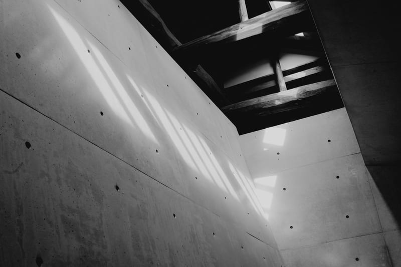 Ahsap kirislerin notr beton duvara dusurdugu sicak gunisigi golgeleri; sade, geometrik mimari detay.
